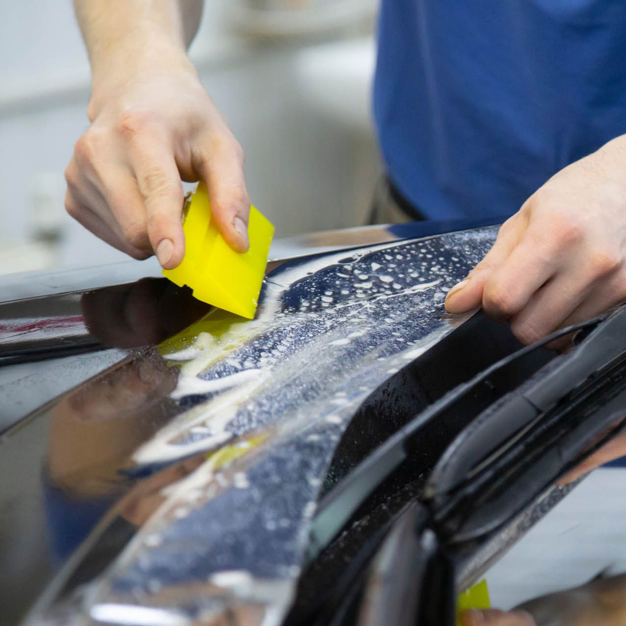Person using a yellow squeegee to apply film to a car's surface, likely for tinting.