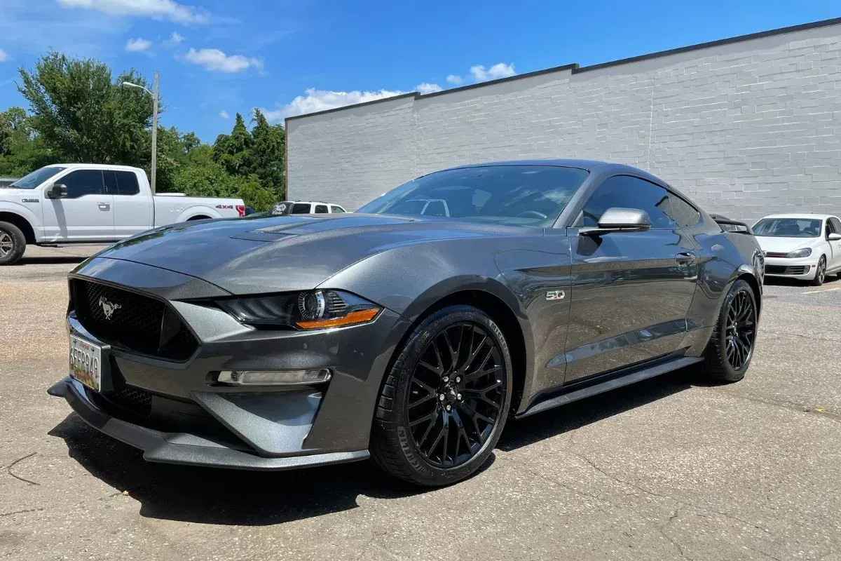 Gray Ford Mustang sports car parked on pavement, with a white truck and building in background.