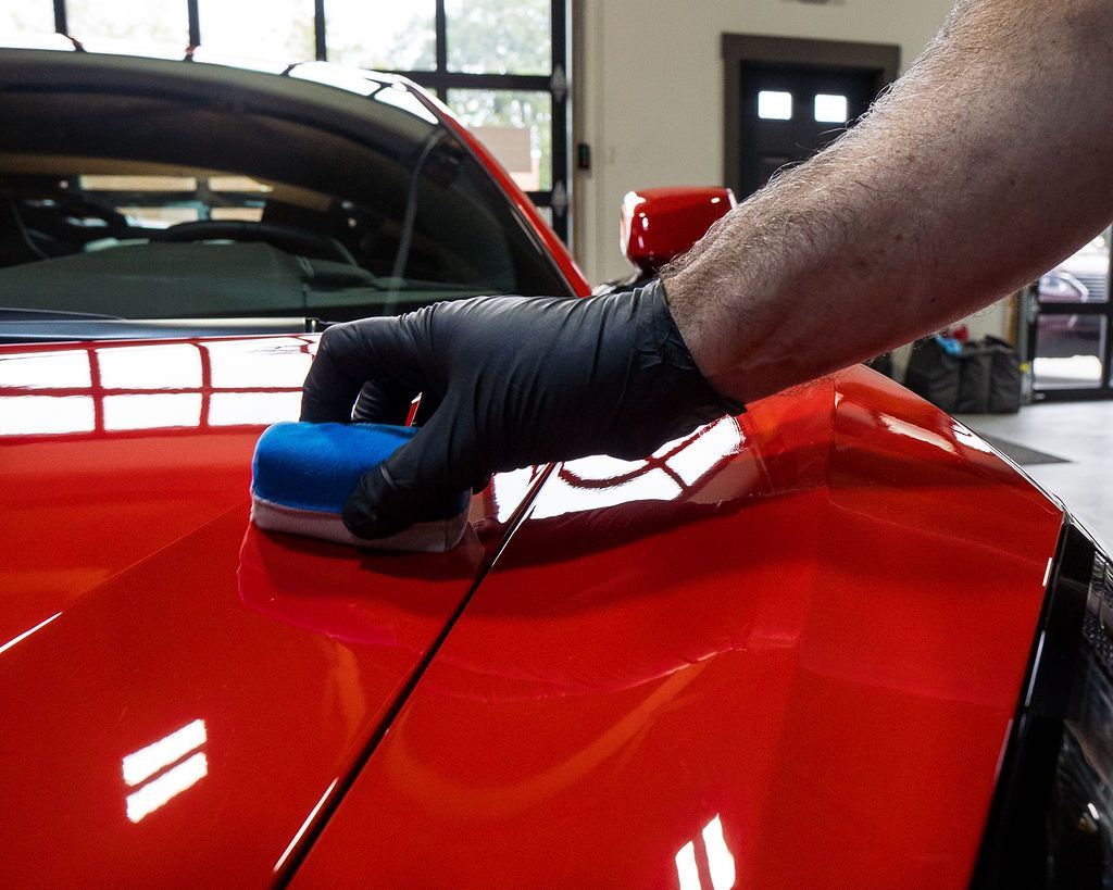 Hand with black glove applying product with blue applicator to red car hood.