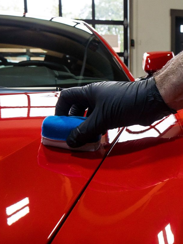 Gloved hand applying ceramic coating to a red car; detail shot of the shiny, reflective paint.