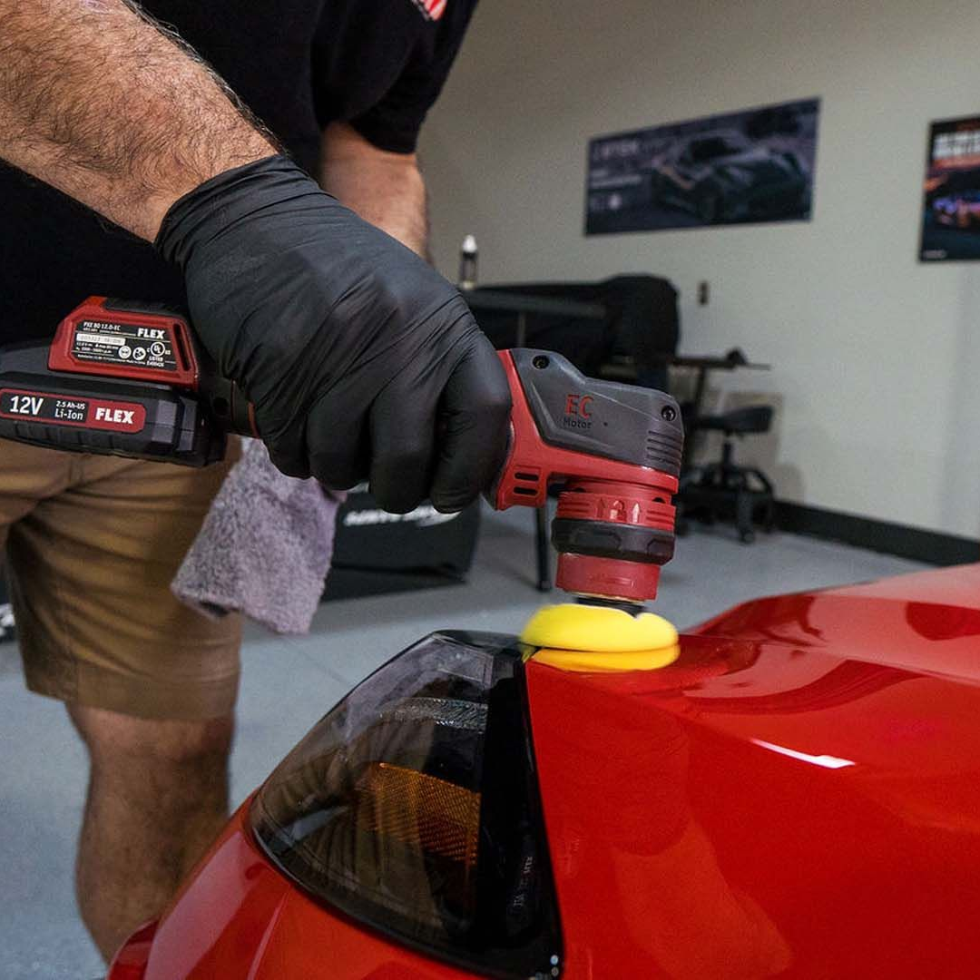 Person polishing a red car with a cordless buffer in a garage, wearing black gloves.