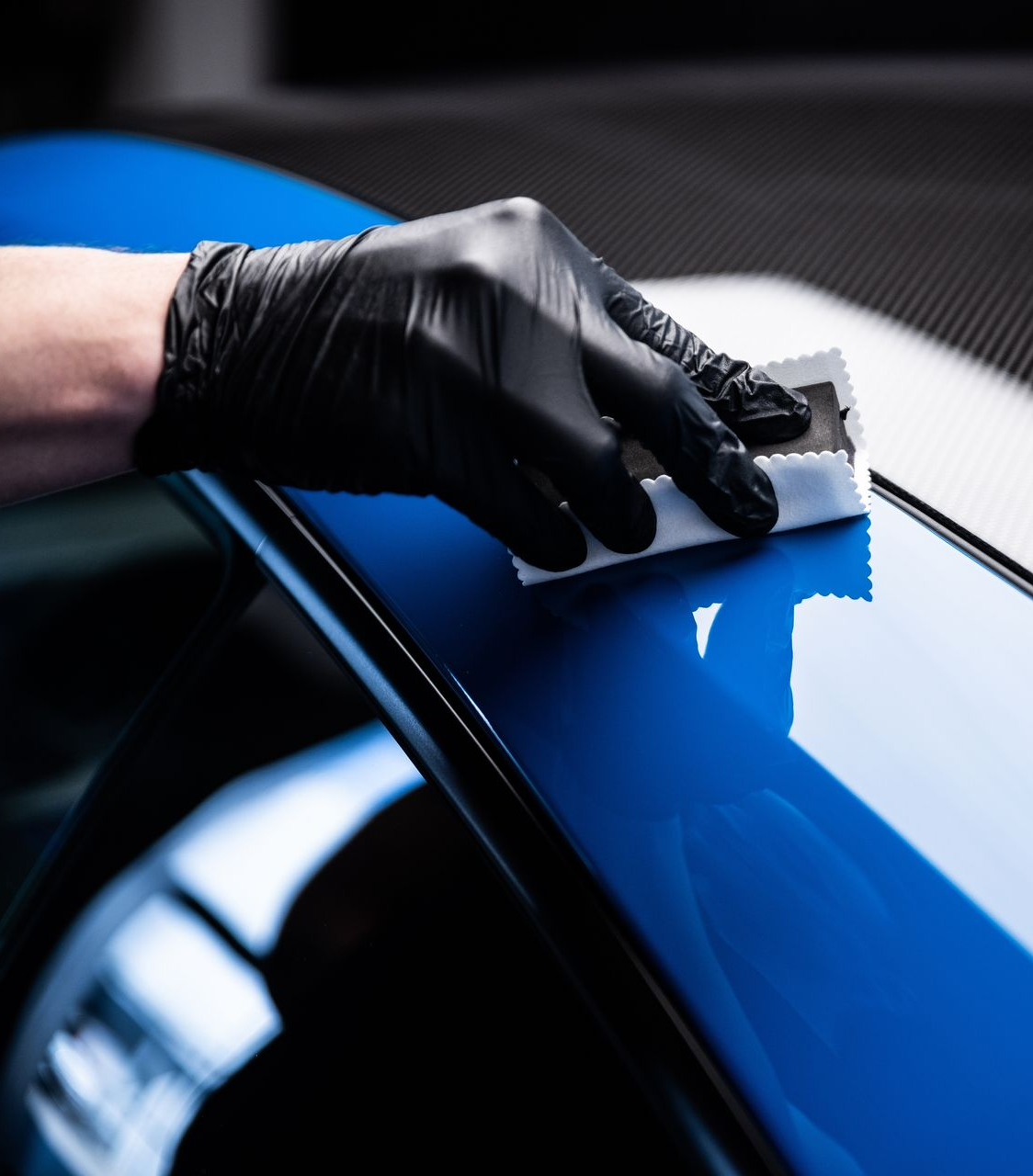 Person in black glove applying ceramic coating to a shiny blue car surface.