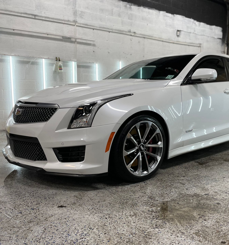 White Cadillac ATS-V sports car in a garage, parked on a reflective floor, under bright lights.