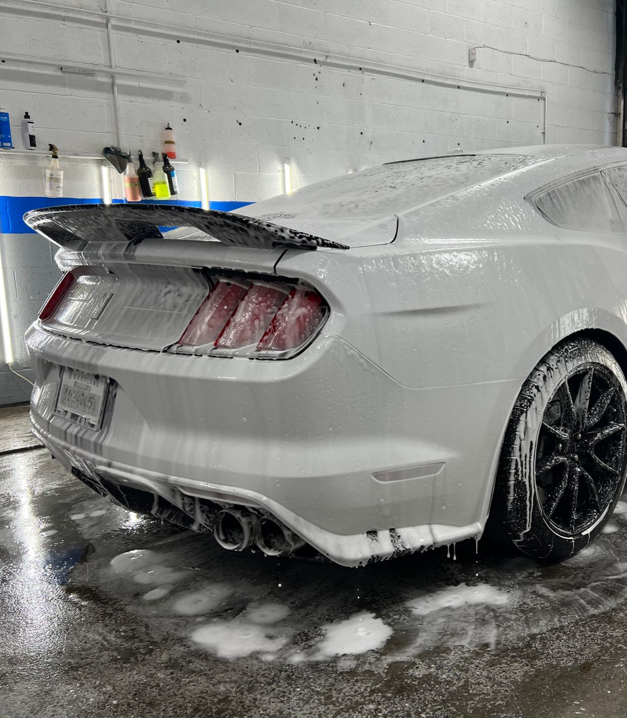 White Ford Mustang covered in soap suds at a car wash, rear view.