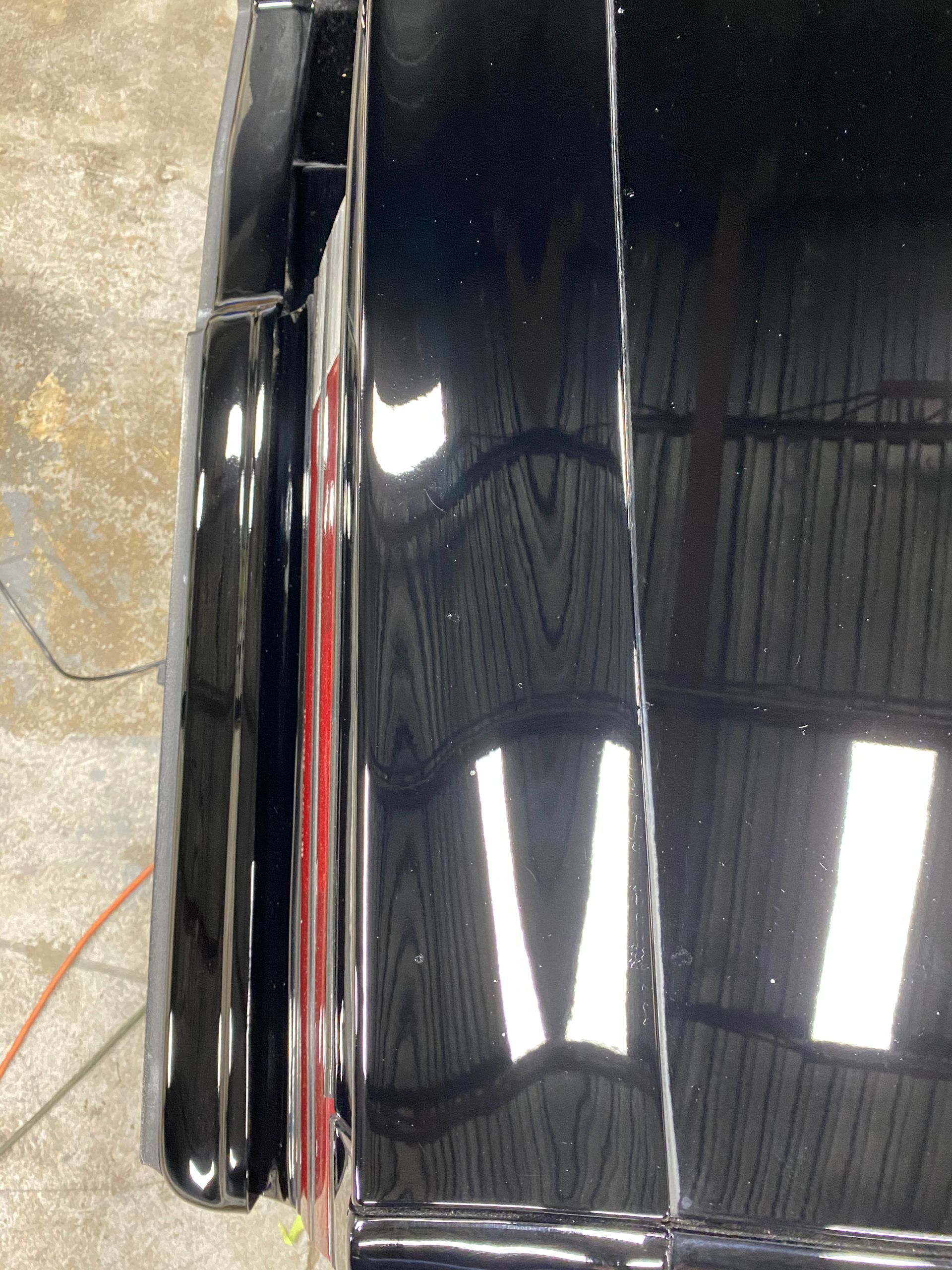 Close-up of a shiny black car fender and bumper. Red strip is visible. Reflective, indoor setting.