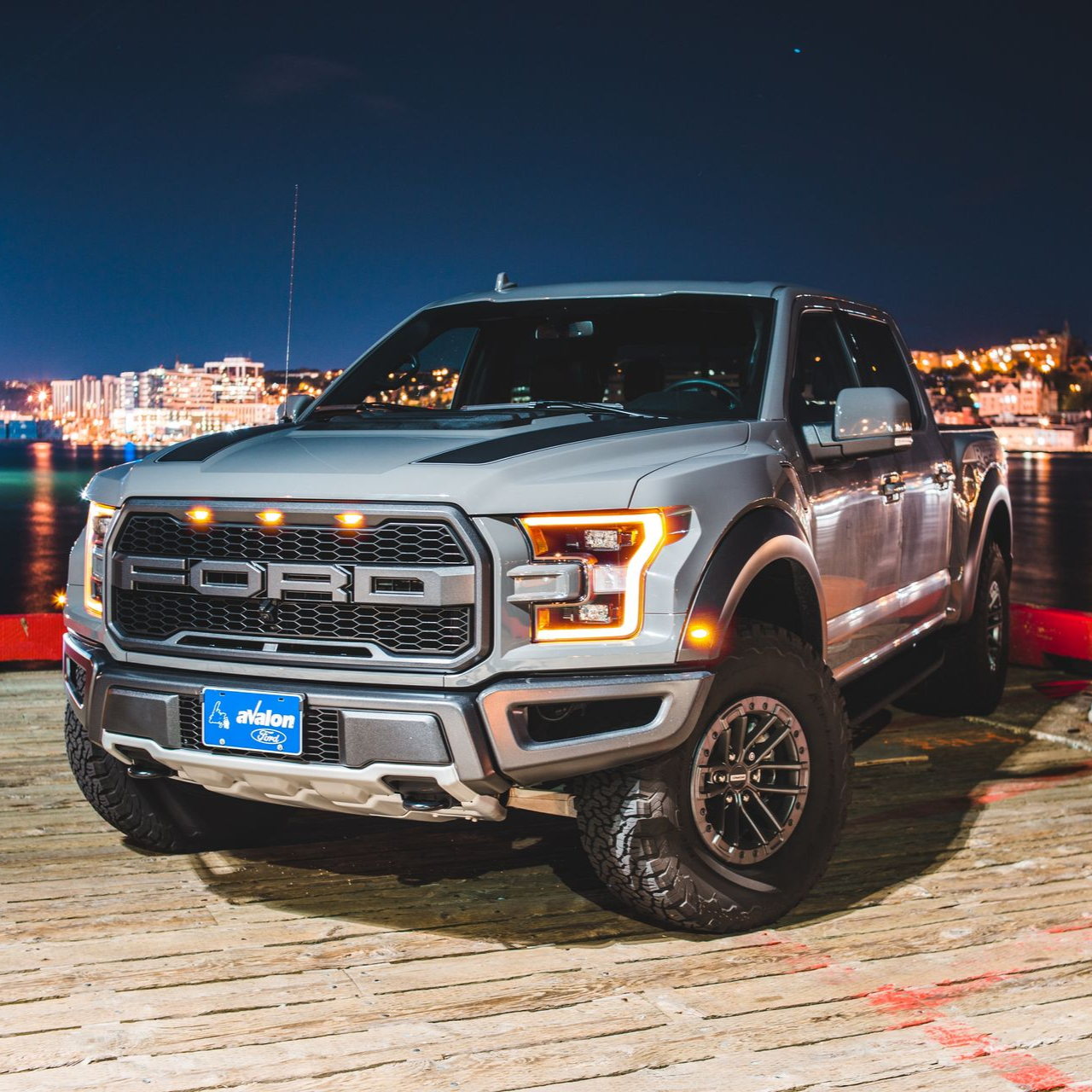 Gray Ford Raptor truck parked on a wooden dock at night with city lights in the background.