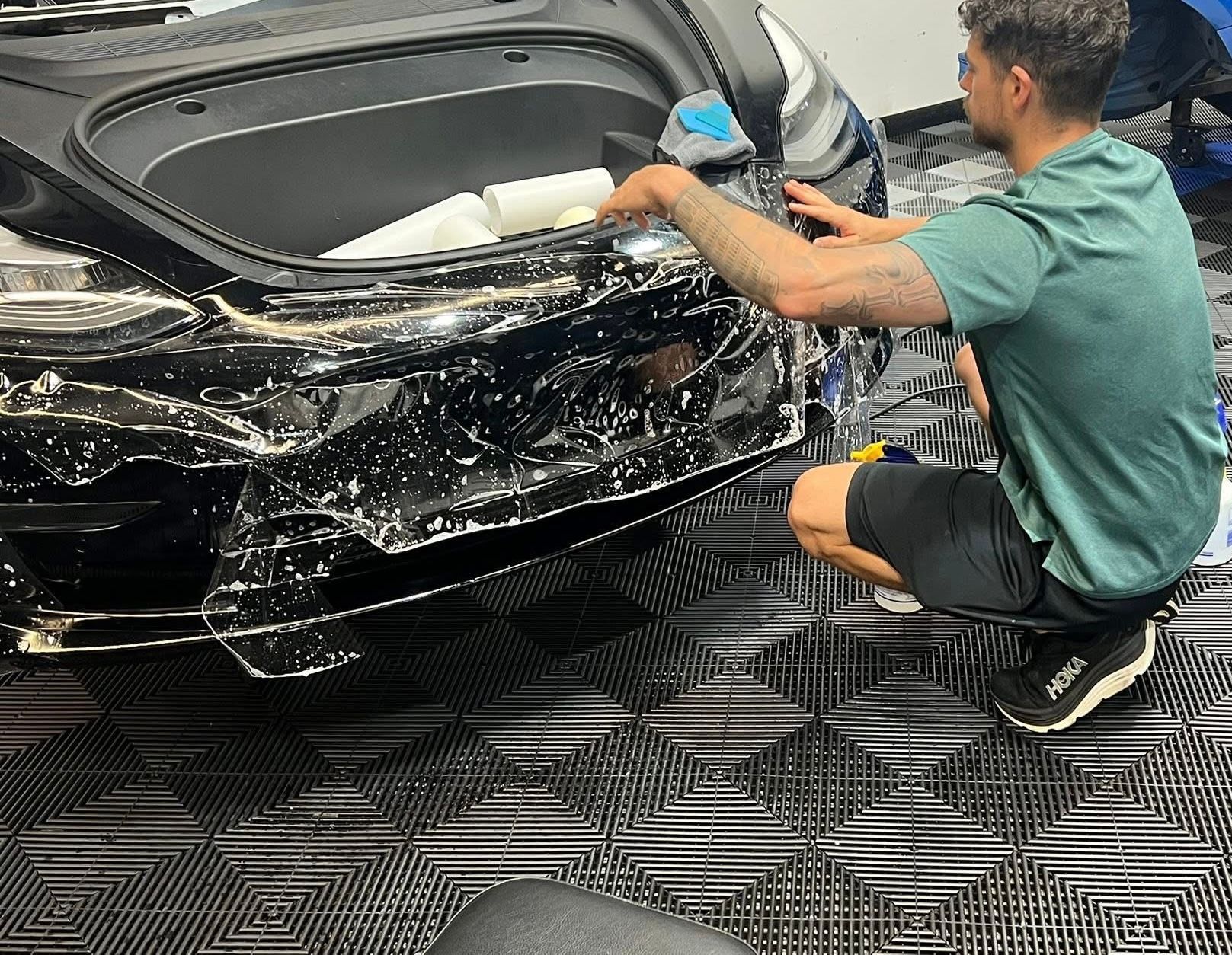 Man applying protective film to a black car bumper in a shop. He's kneeling, wearing shorts, and a t-shirt.