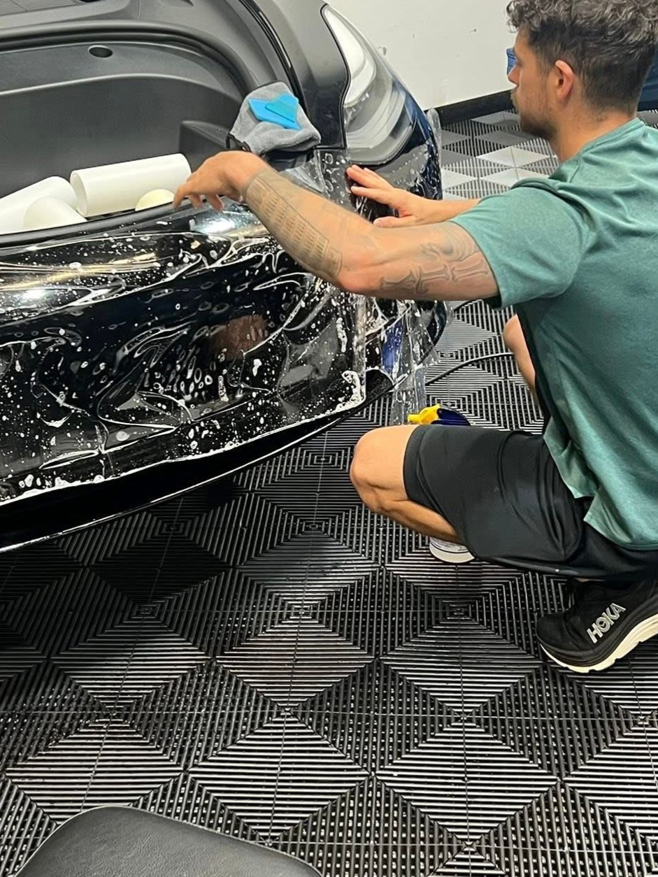 Man installing a protective film on a black car bumper in a garage. He squats, wiping.