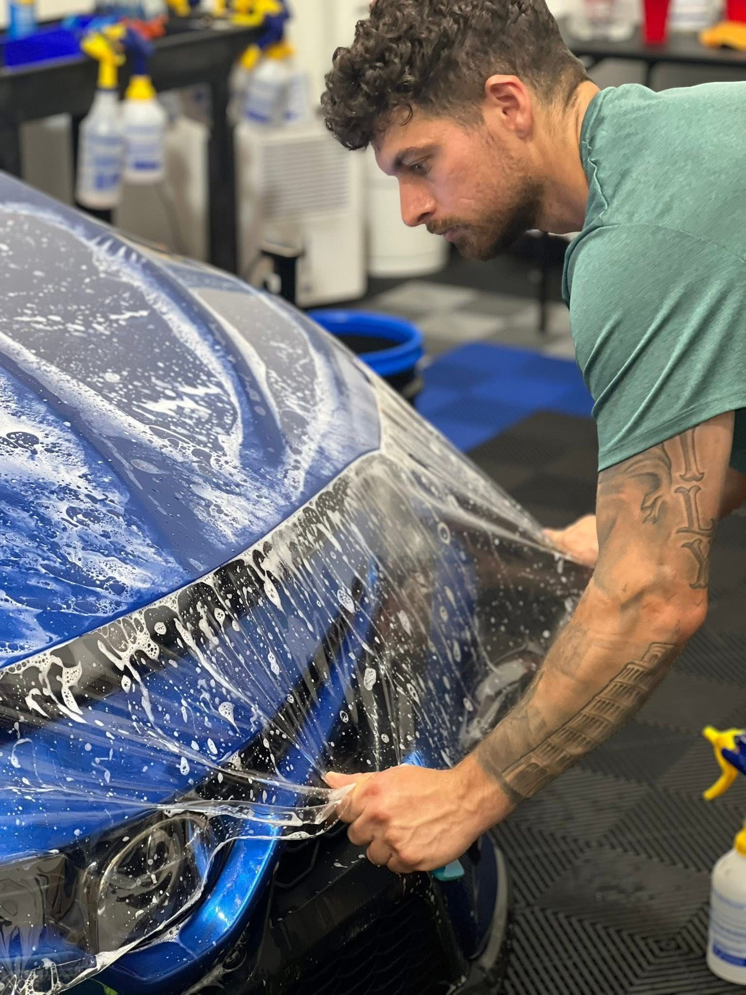 Man applying protective film to a blue car in a workshop.