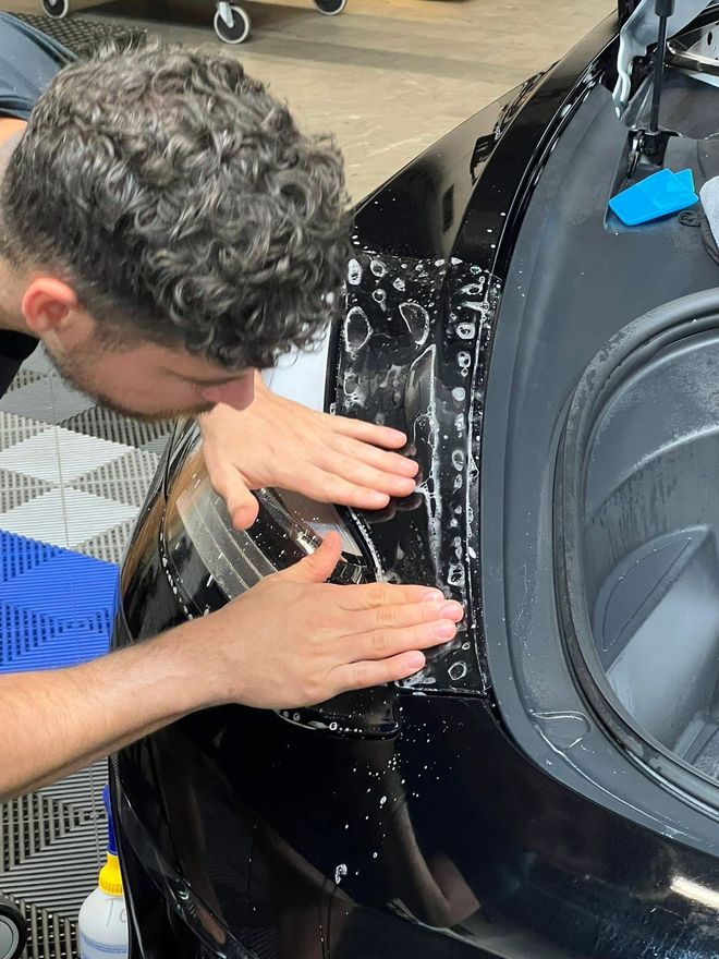 Man applying film to a black car headlight; indoors. Hands pressing, focused expression.