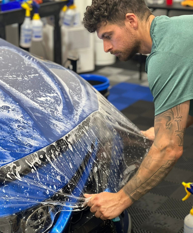 Man applying protective film to a blue car in a garage.