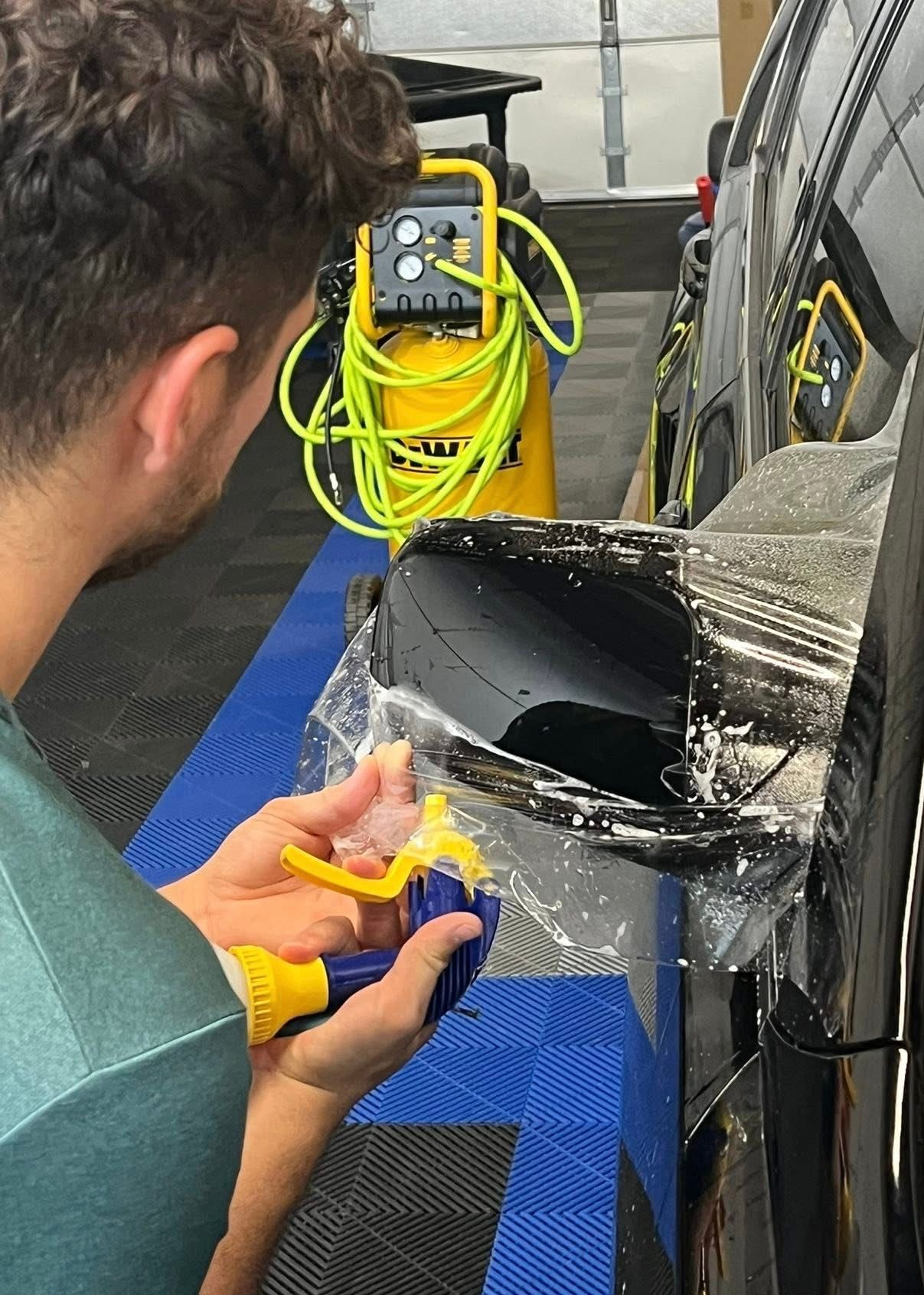 Man spraying water on a car mirror wrapped in clear film. Garage setting, yellow/blue spray bottle.