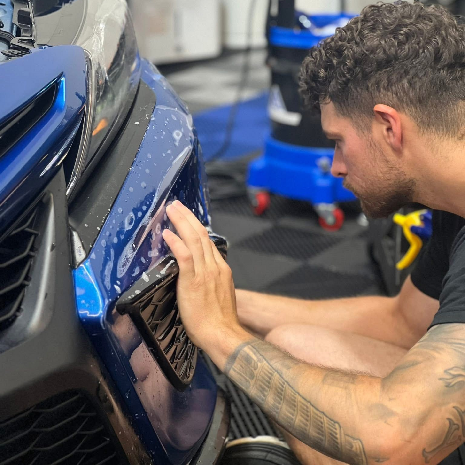 Man installing protective film on a blue car bumper in a garage.