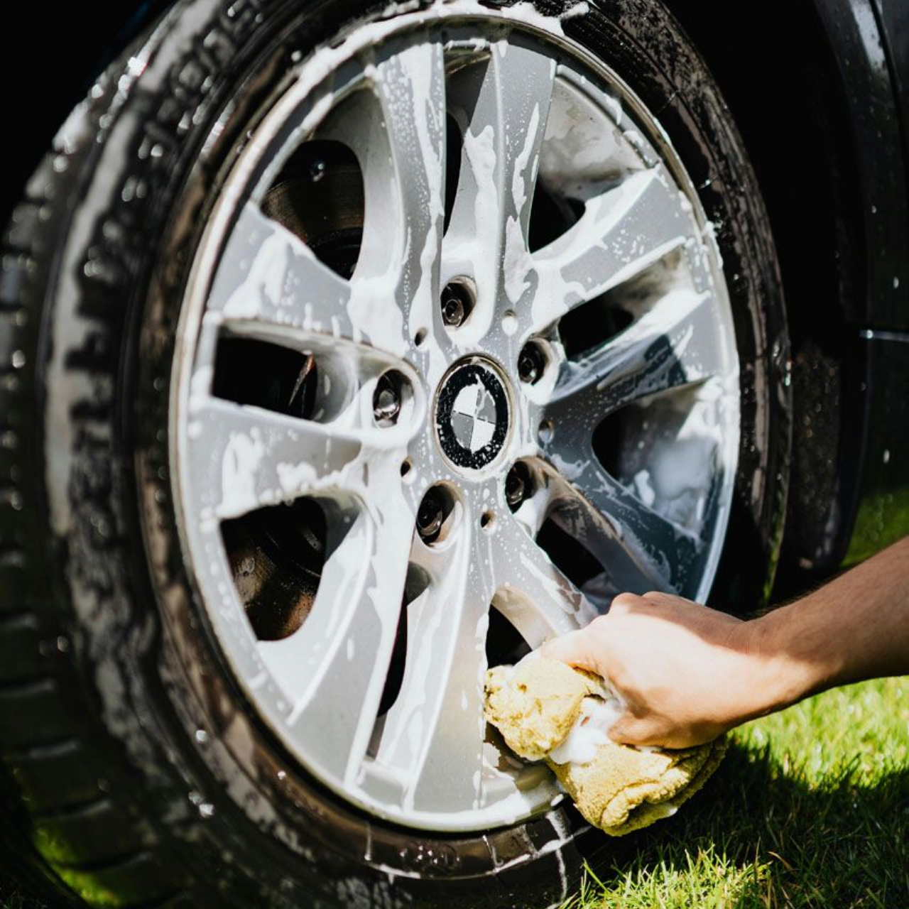 Person washing a car tire with soapy water and a sponge; outdoors on grass.