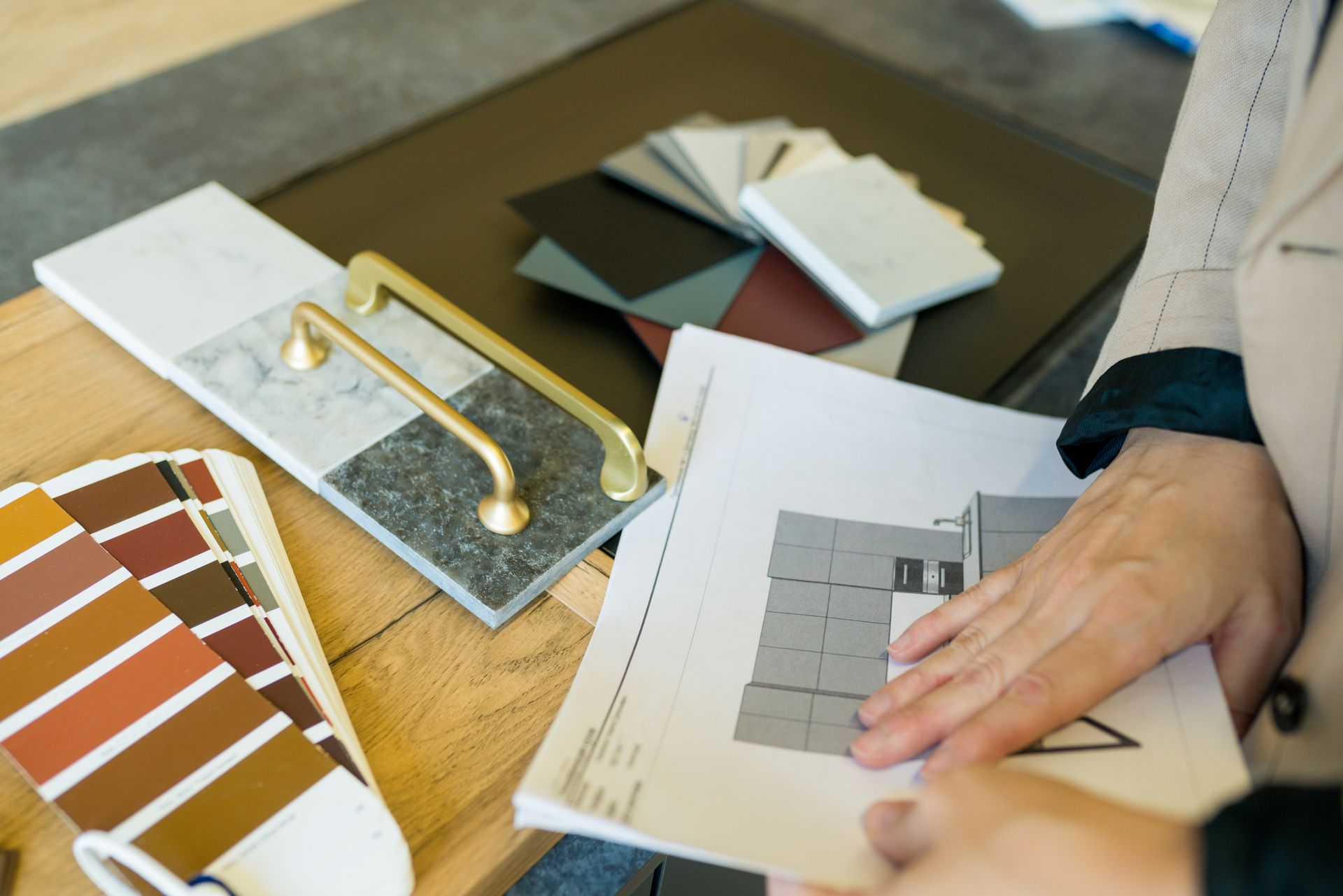 Person holding architectural plans with colour swatches, tile samples, and cabinet hardware on a table.