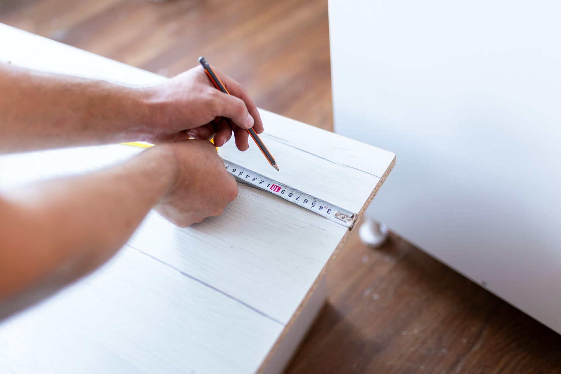 Builder using a measuring tape and pencil to mark measurements on a white wooden board.