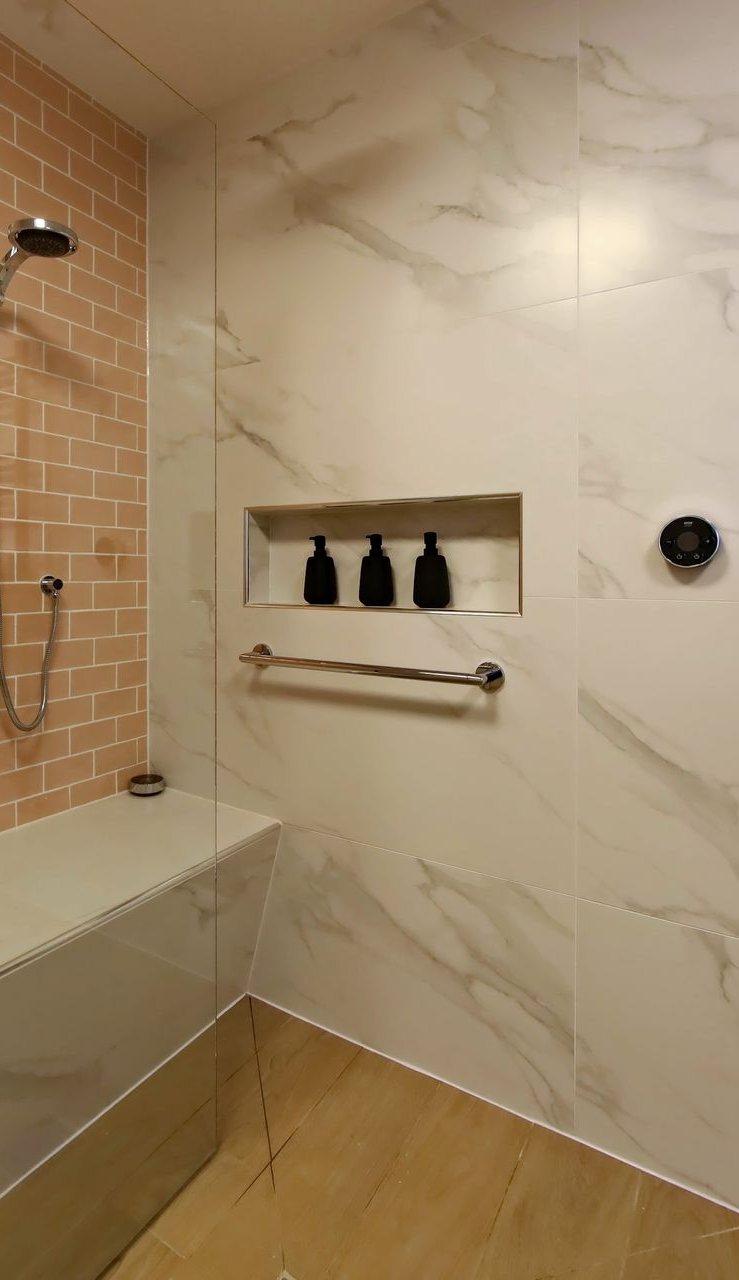 Bathroom shower with white marble-look tile walls, built-in shelving with black bottles, pink brick wall, and chrome fixtures.