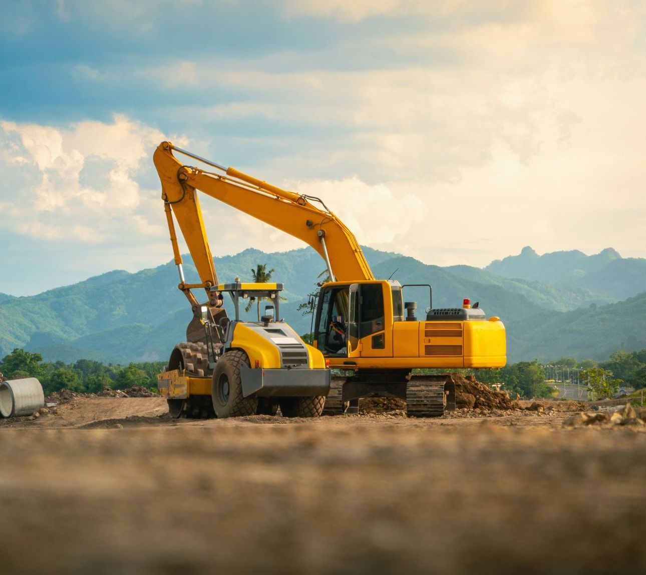 A yellow excavator is working on a construction site with mountains in the background.