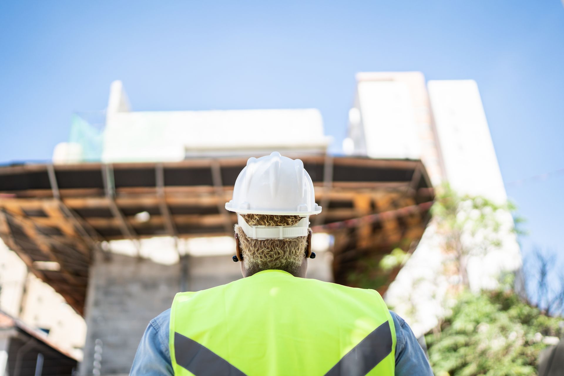 A construction worker wearing a hard hat and safety vest is looking at a building under construction.
