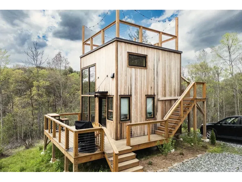 Wooden cabin with deck and rooftop terrace in a wooded area; black car parked nearby.