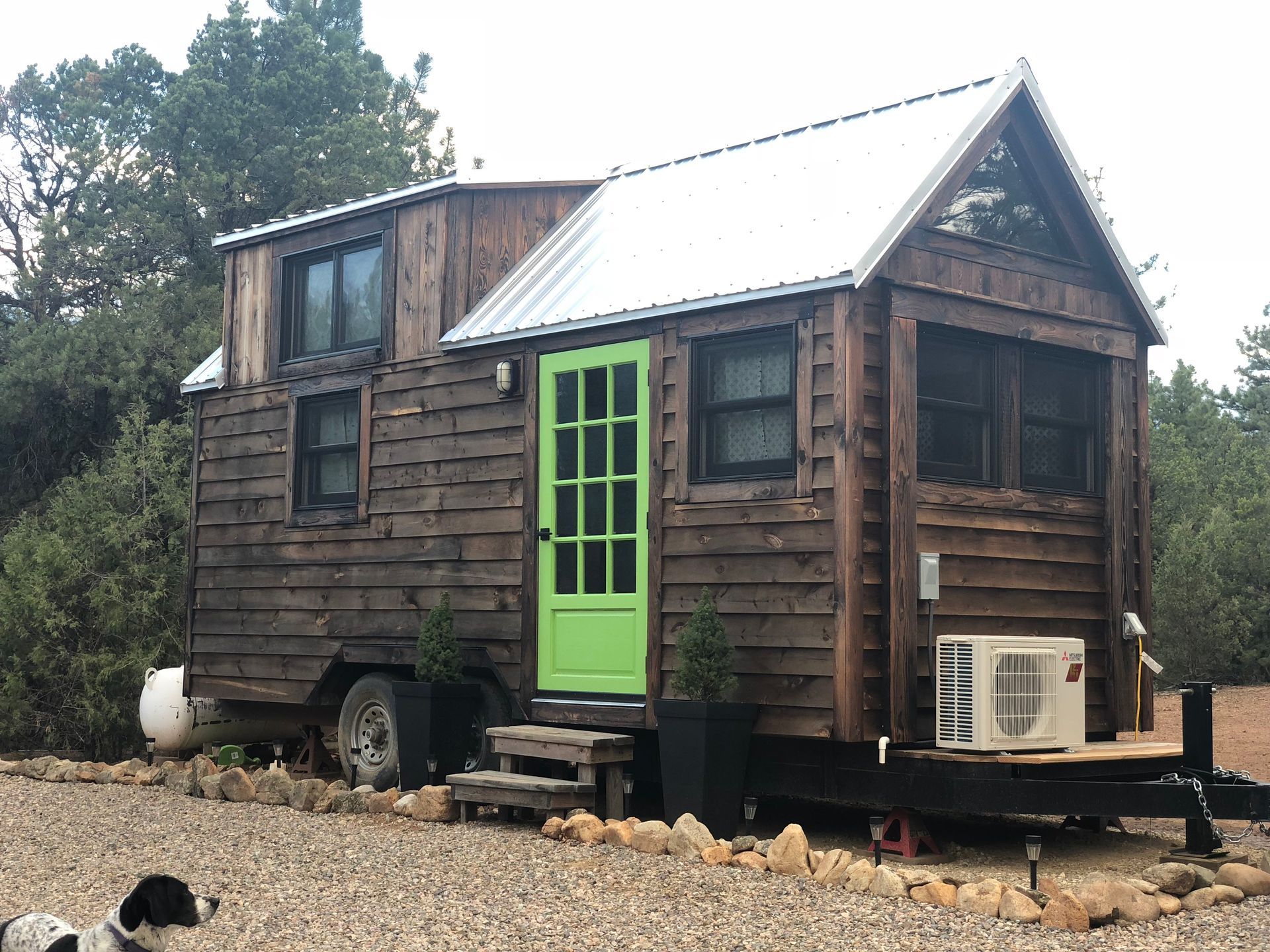 Rustic tiny home on wheels with wood siding, green door, and loft windows, parked in a wooded area.