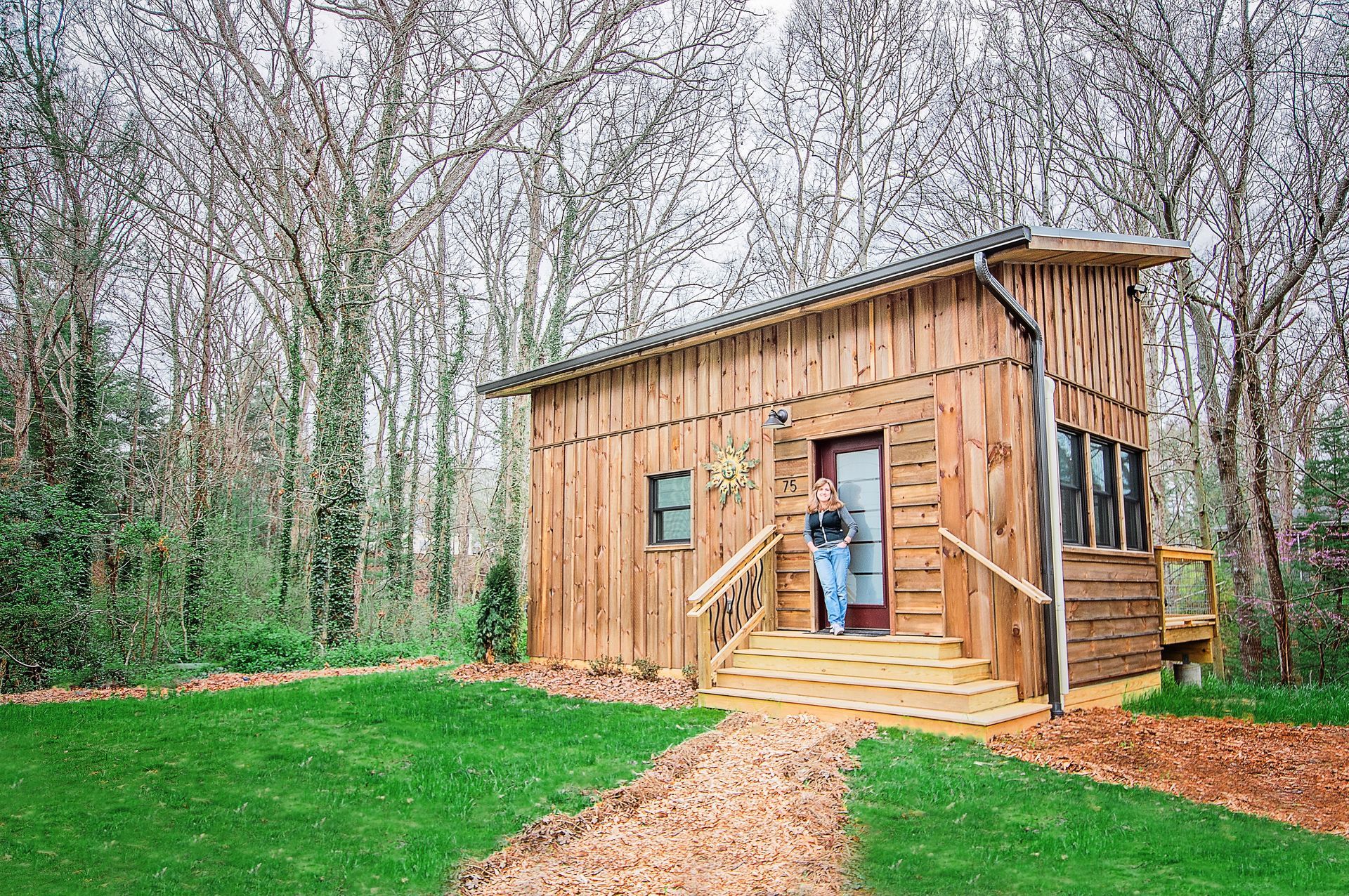 A woman is standing in front of a tiny wooden home in the woods.