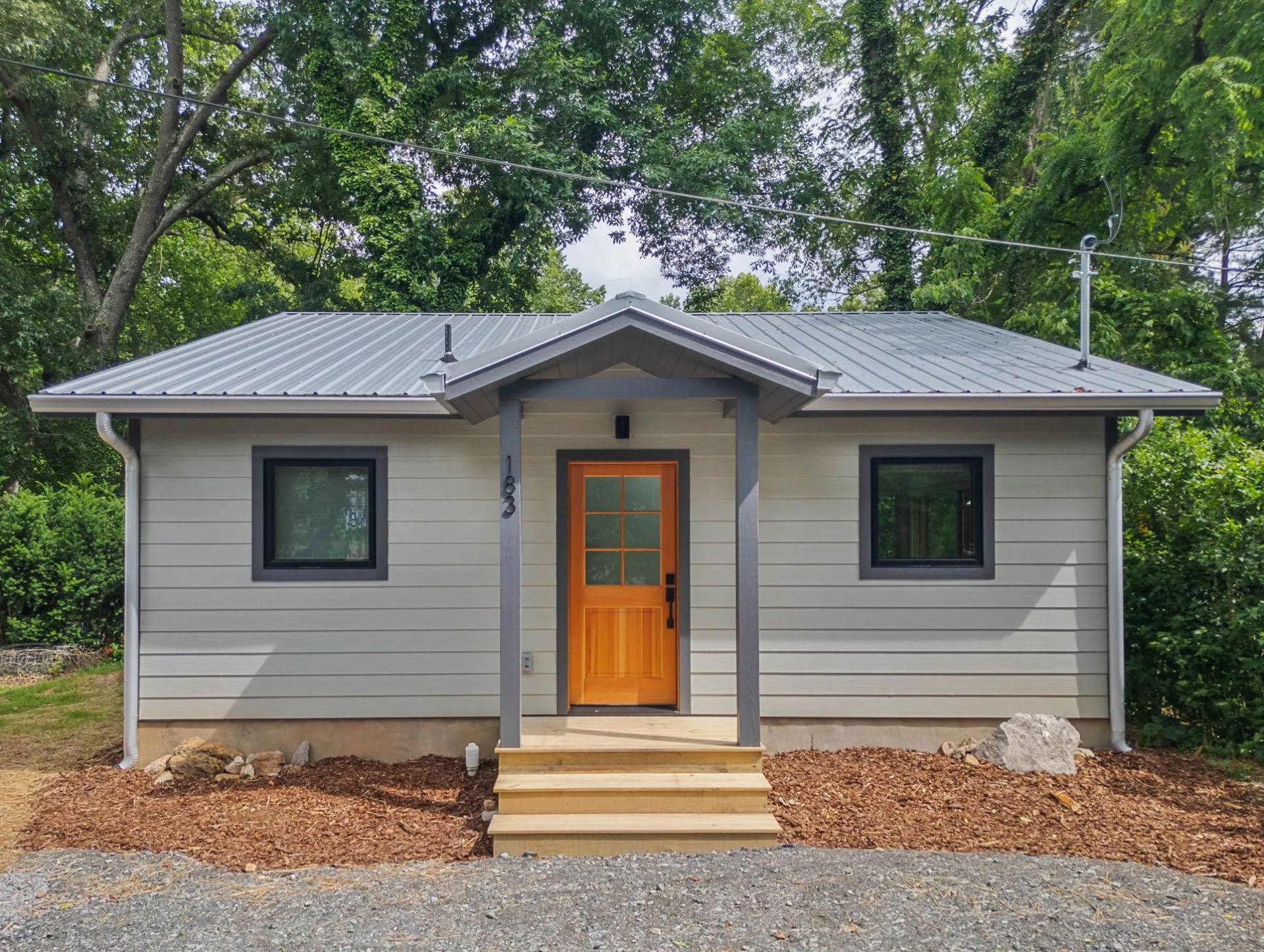 A modern, compact home with light gray siding, a metal roof, and a bright orange front door, surrounded by lush greenery and trees.