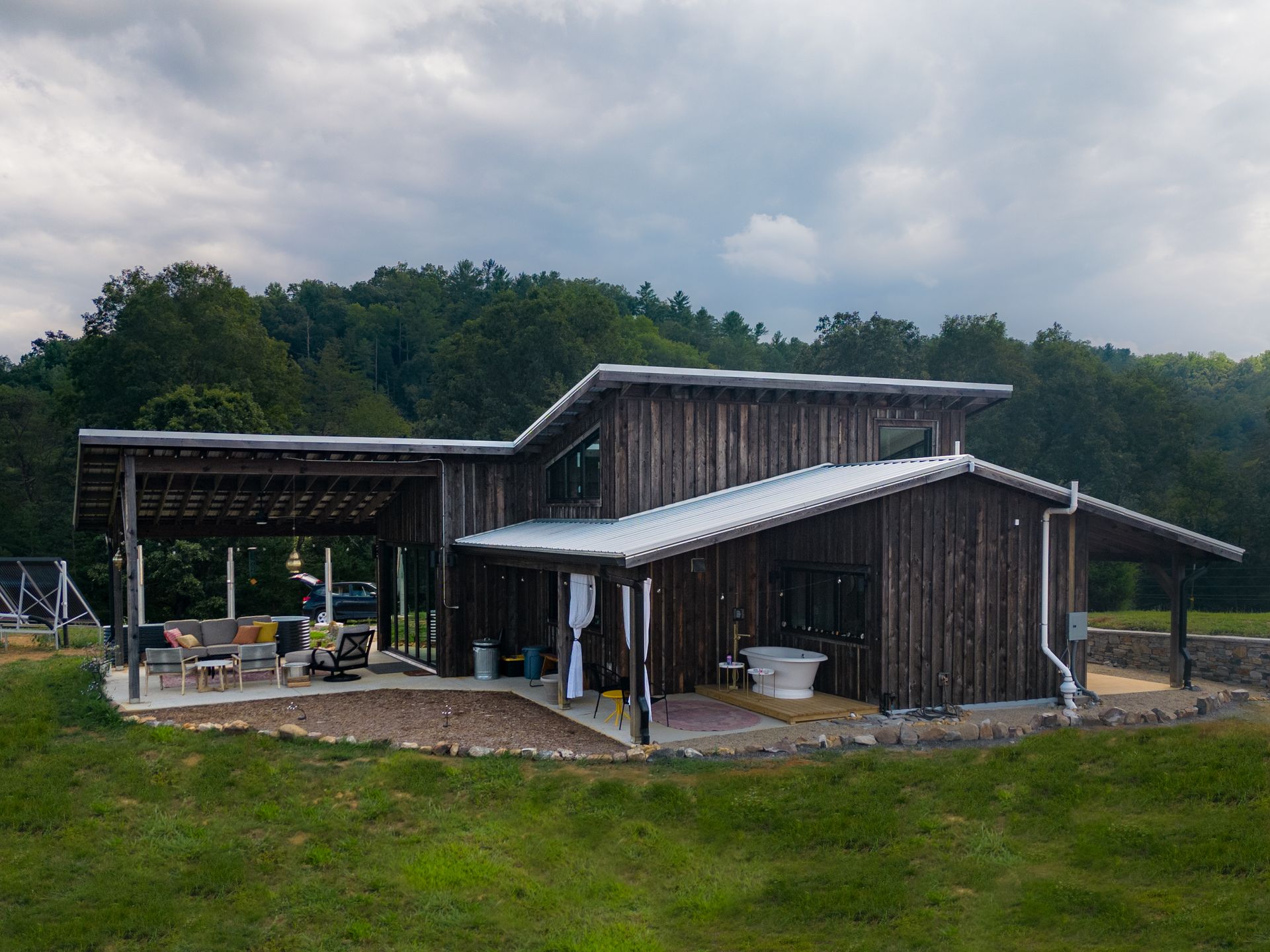 Modern rustic house with sloped metal roofs and wood siding, set on a grassy lot with forested hills