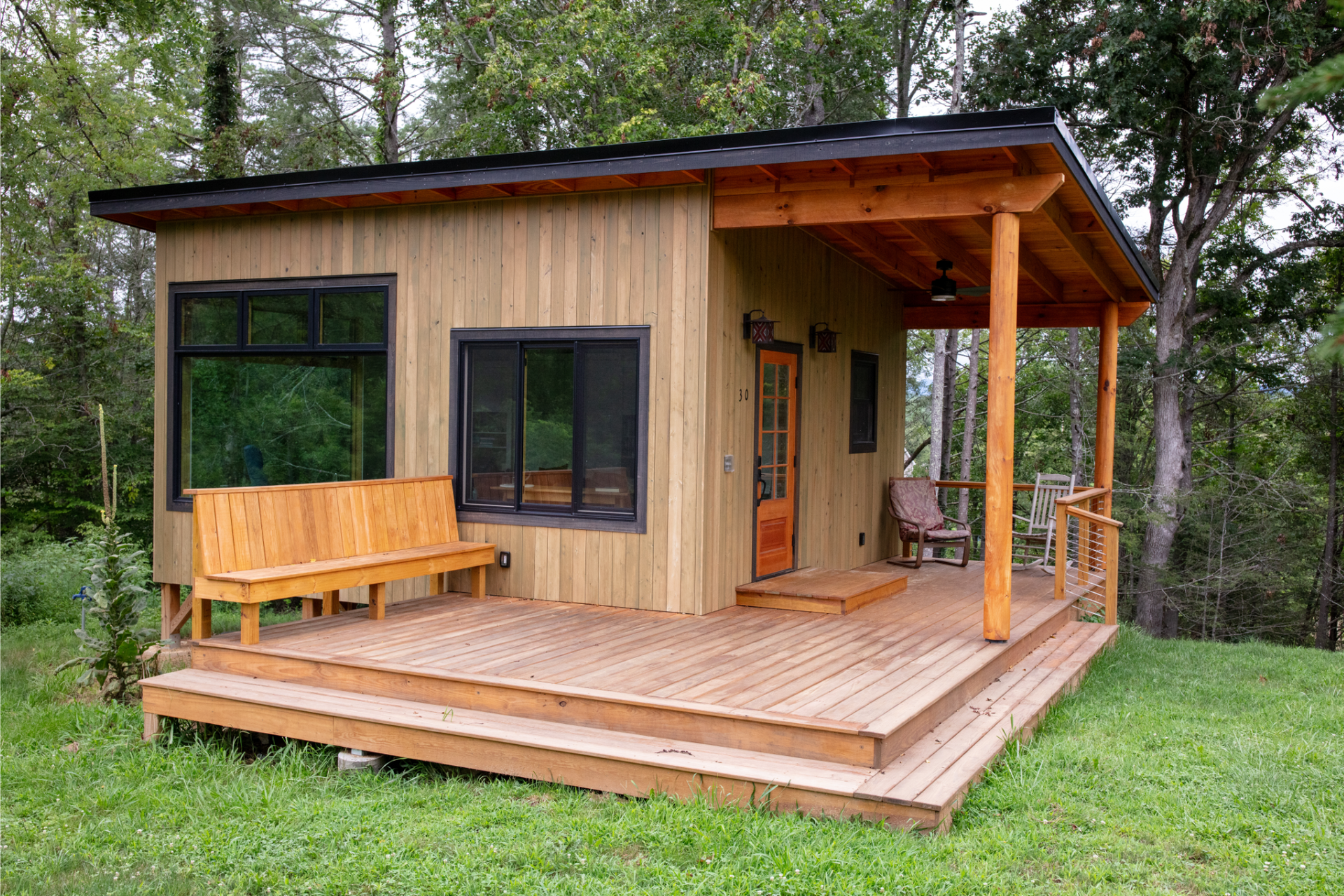 A small, modern wooden cabin with a deck, large windows, and a covered porch area set in a green, wooded landscape.