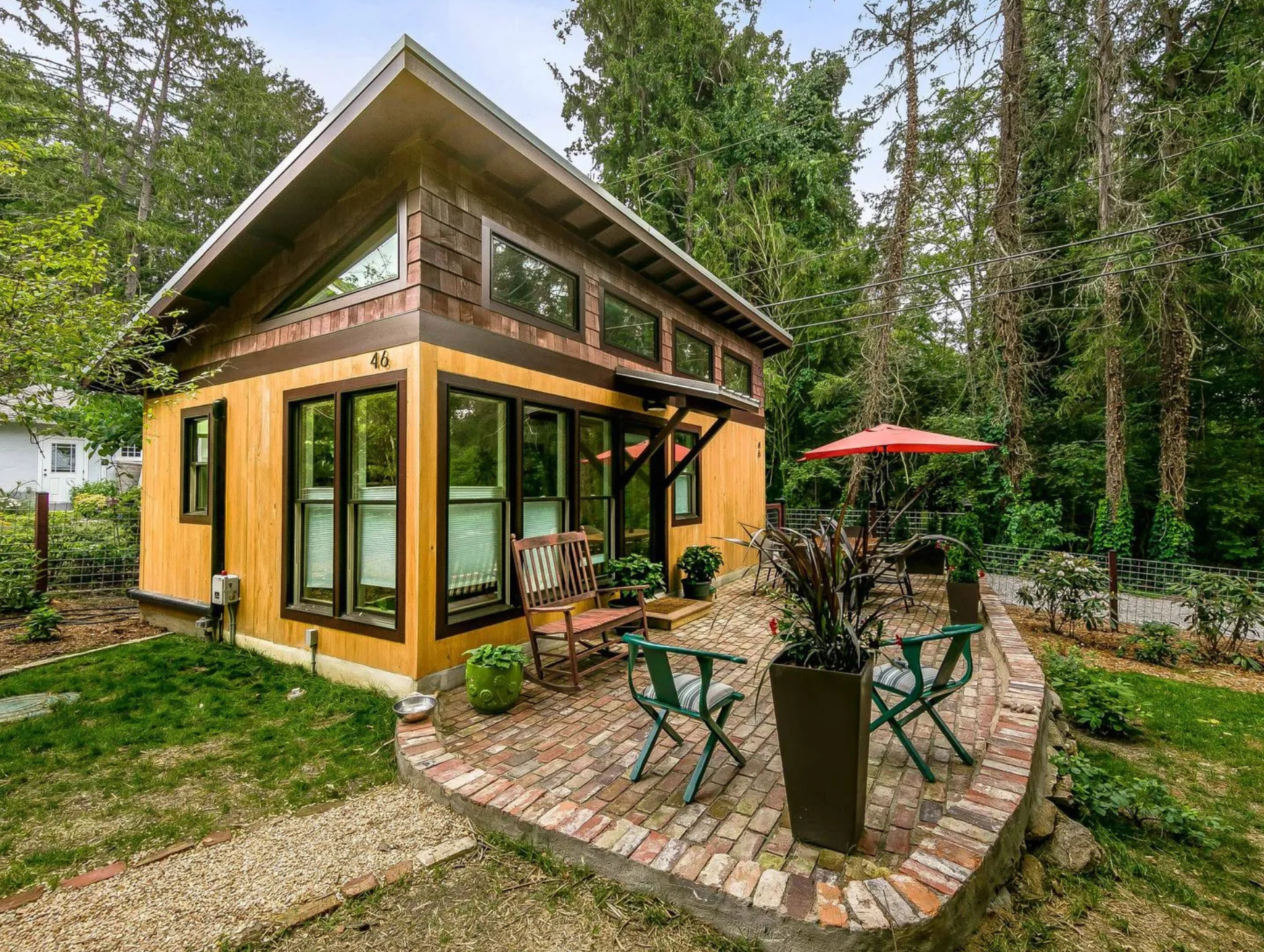 A modern wood-sided cabin with large windows and a slanted roof sits on a brick patio with patio chairs and an umbrella.