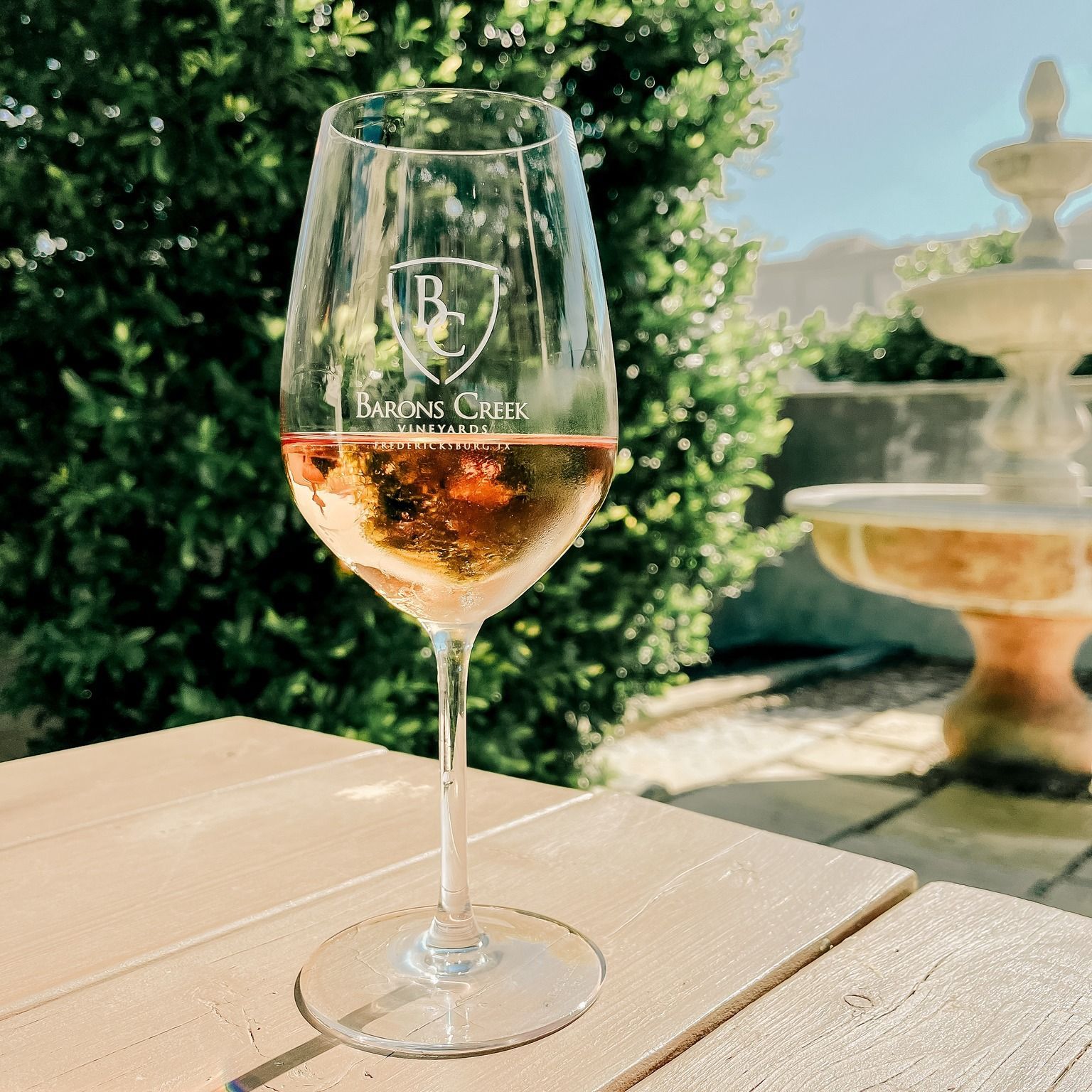 A wine glass filled with rosé on a wooden table outdoors, with a fountain and green bush in the background.