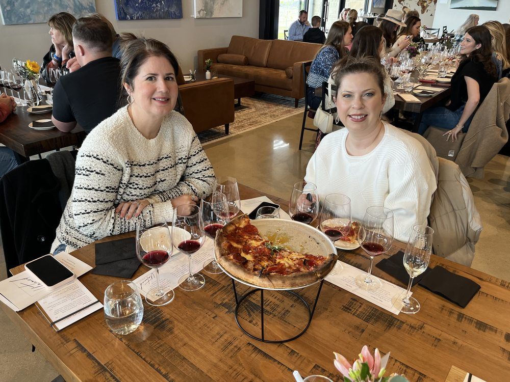 Two women smiling at a table with pizza and wine in a restaurant. People in the background.