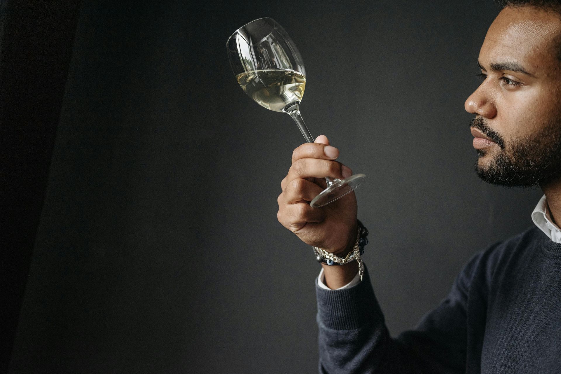 Man holding up a glass of white wine, examining it closely against a dark background.