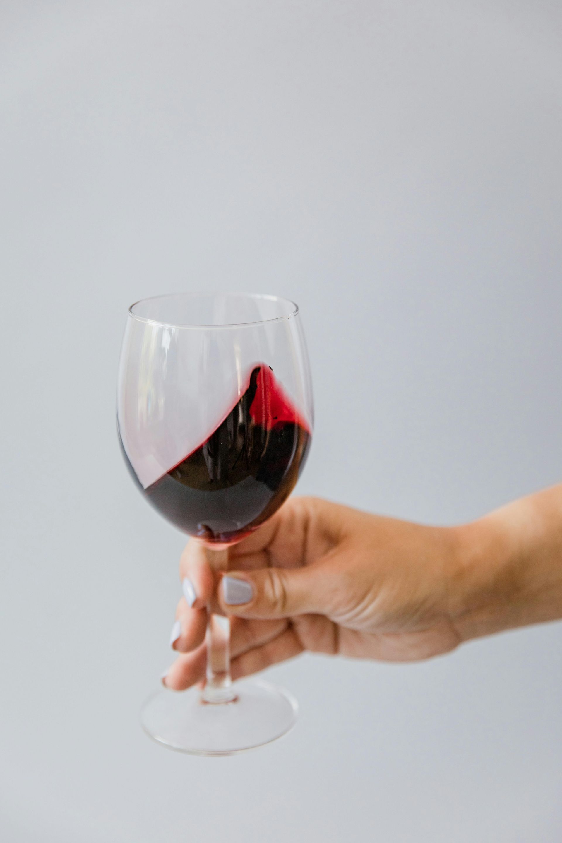 Hand holding a wine glass filled with red wine, swirling against a gray backdrop.