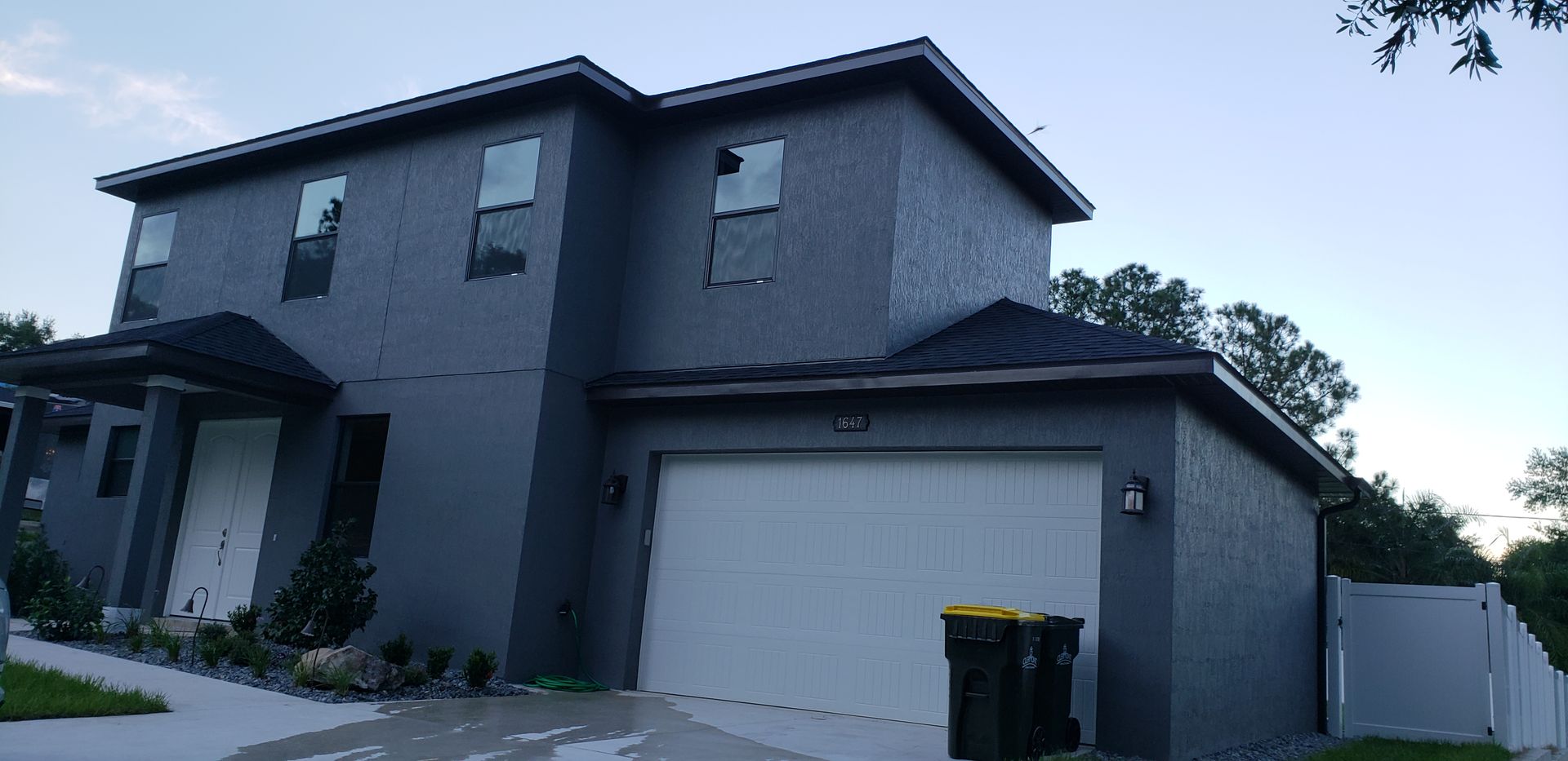 A large gray house with a white garage door and a trash can in front of it.
