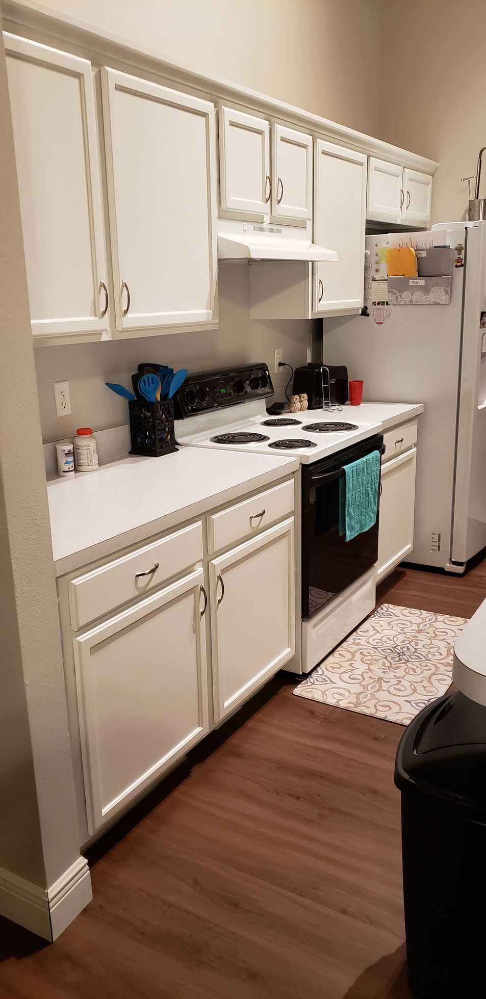 A kitchen with white cabinets , a stove , a refrigerator , and a trash can.