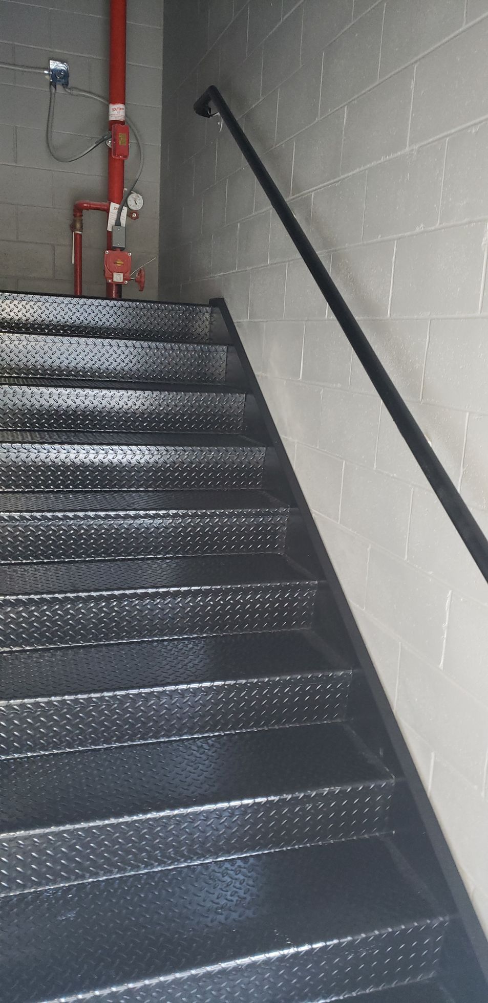 A set of black stairs with a black railing and a red pipe on the wall.