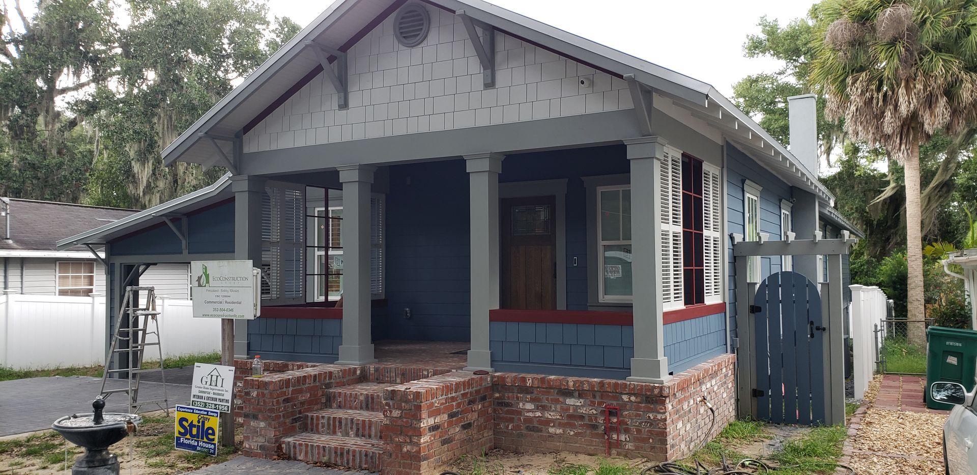 A small blue and white house with a porch and a fence in front of it.