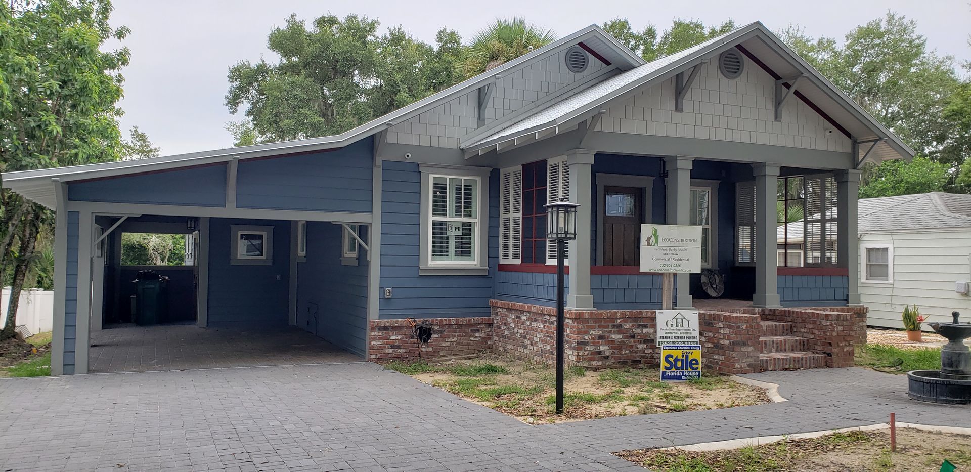 A blue and white house with a carport and a sign in front of it.