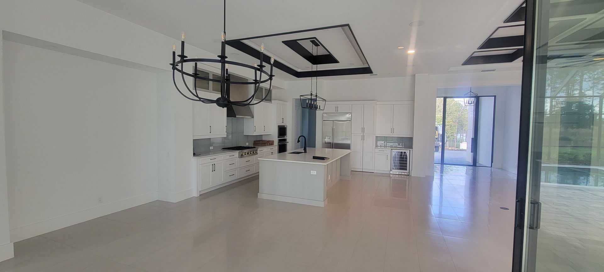 A large empty room with a kitchen and a chandelier hanging from the ceiling.