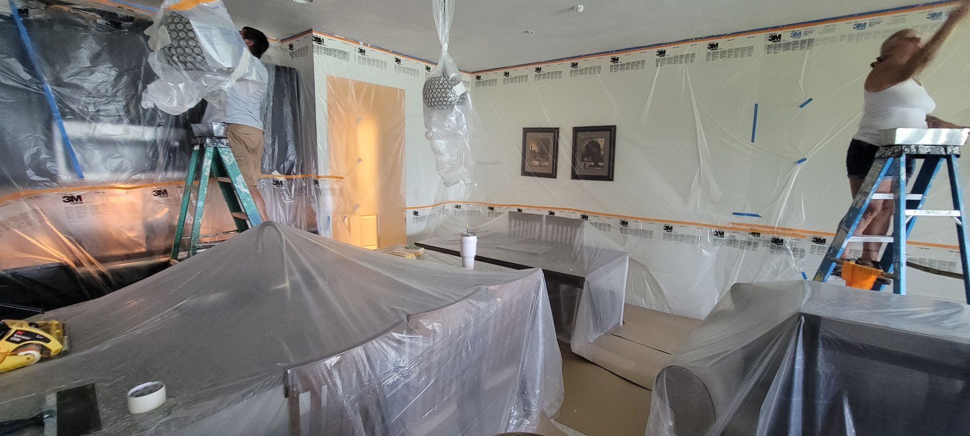 A couple of people are standing on ladders in a living room covered in plastic.
