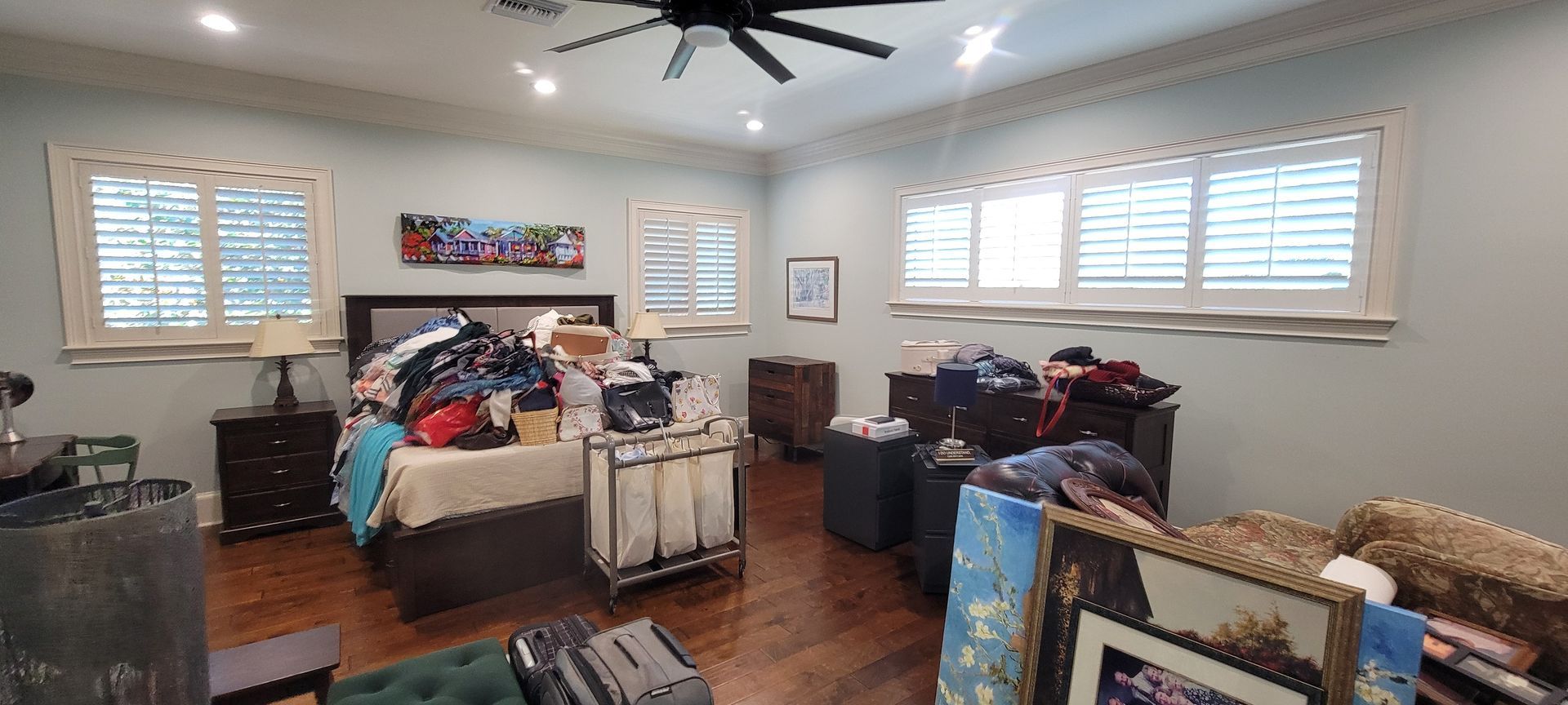A bedroom with a ceiling fan , a bed , a couch , and a lot of clothes.