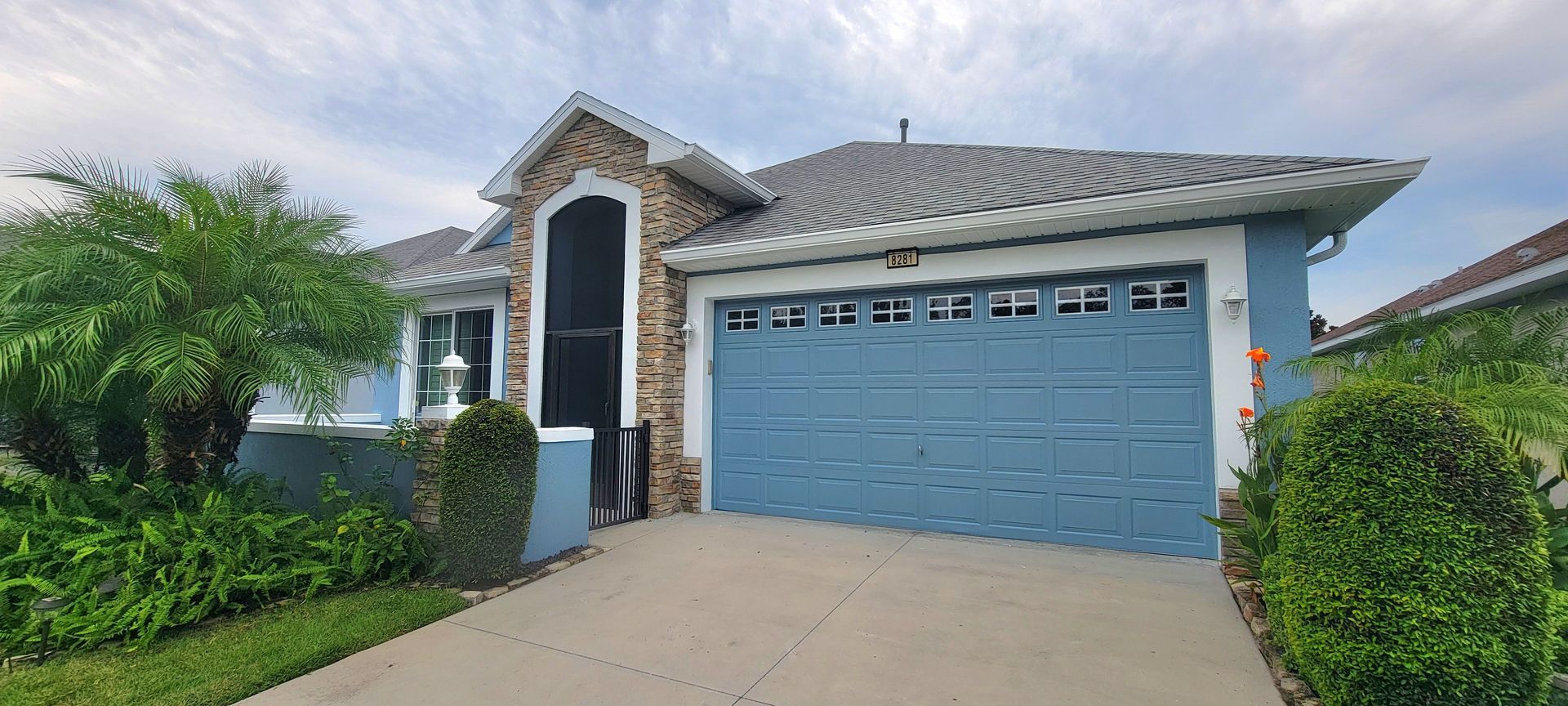 A house with a blue garage door, a stone facade, and landscaping under a cloudy sky.