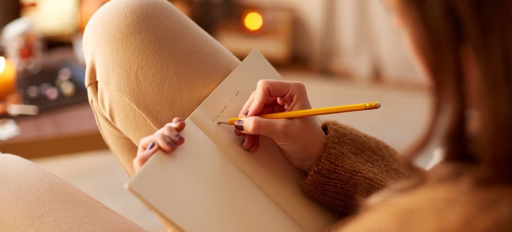 Person writes in a notebook with a pencil, legs crossed, cozy setting.