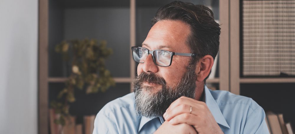 Man with glasses and a beard looking off-camera, resting hands on each other.