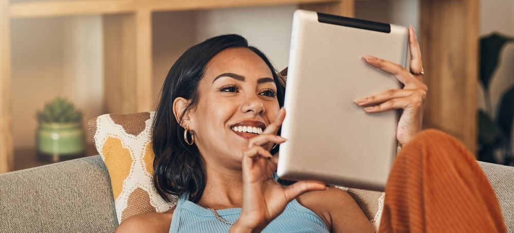 Woman smiles while using a tablet, relaxed on a couch. Indoors, warm lighting.