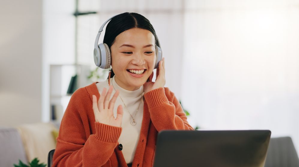 Woman in headphones smiles and waves at laptop screen in a brightly lit room.