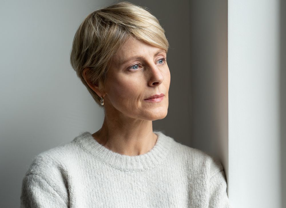 Woman with short blonde hair, looking out a window with a pensive expression. She wears a cream sweater.