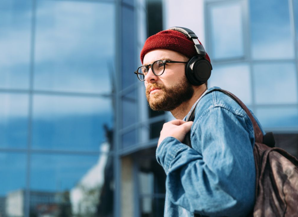 Man with glasses, headphones, and backpack near a building with blue glass. He's wearing a red beanie and denim jacket.
