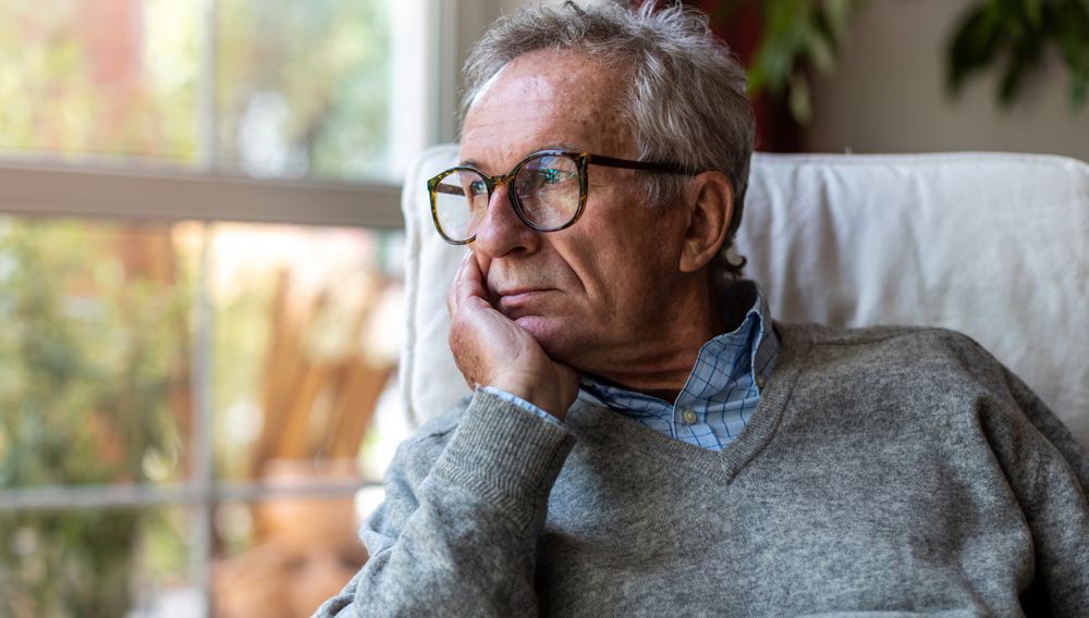 Man in glasses, resting his chin on his hand, looking thoughtful near a window.